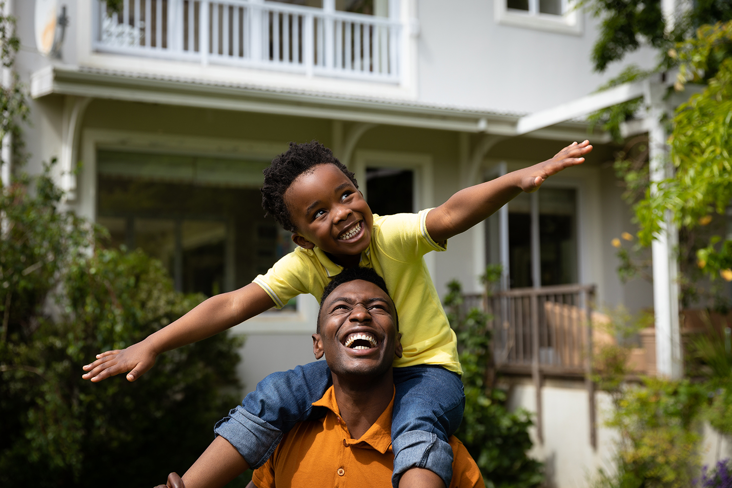 Front view of a smiling African American man in the garden carrying his son piggyback, the boy stretching his arms out